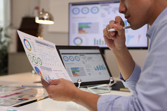 businessperson reading charts and graphs on paper, laptop and a screen
