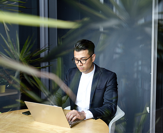 Man sitting on a white chair at a wooden desk in a glass wall office, looking at a laptop